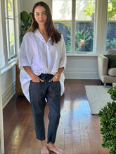 Load image into Gallery viewer, Woman wearing a white shirt and dark pants standing in a sunlit room with large windows.