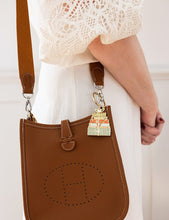 Load image into Gallery viewer, Brown handbag with a decorative charm held by a person wearing a white lace top.