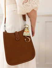 Load image into Gallery viewer, Brown leather handbag with a decorative keychain held by a person wearing a white dress.