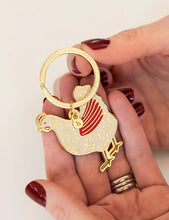 Load image into Gallery viewer, Hand holding a gold and red keychain with a bird design on a beige background